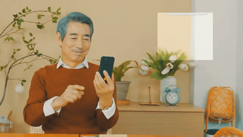 거실에서 중년 남성이 스마트폰 앱 화면을 보며 웃는 해외 홍보영상 장면